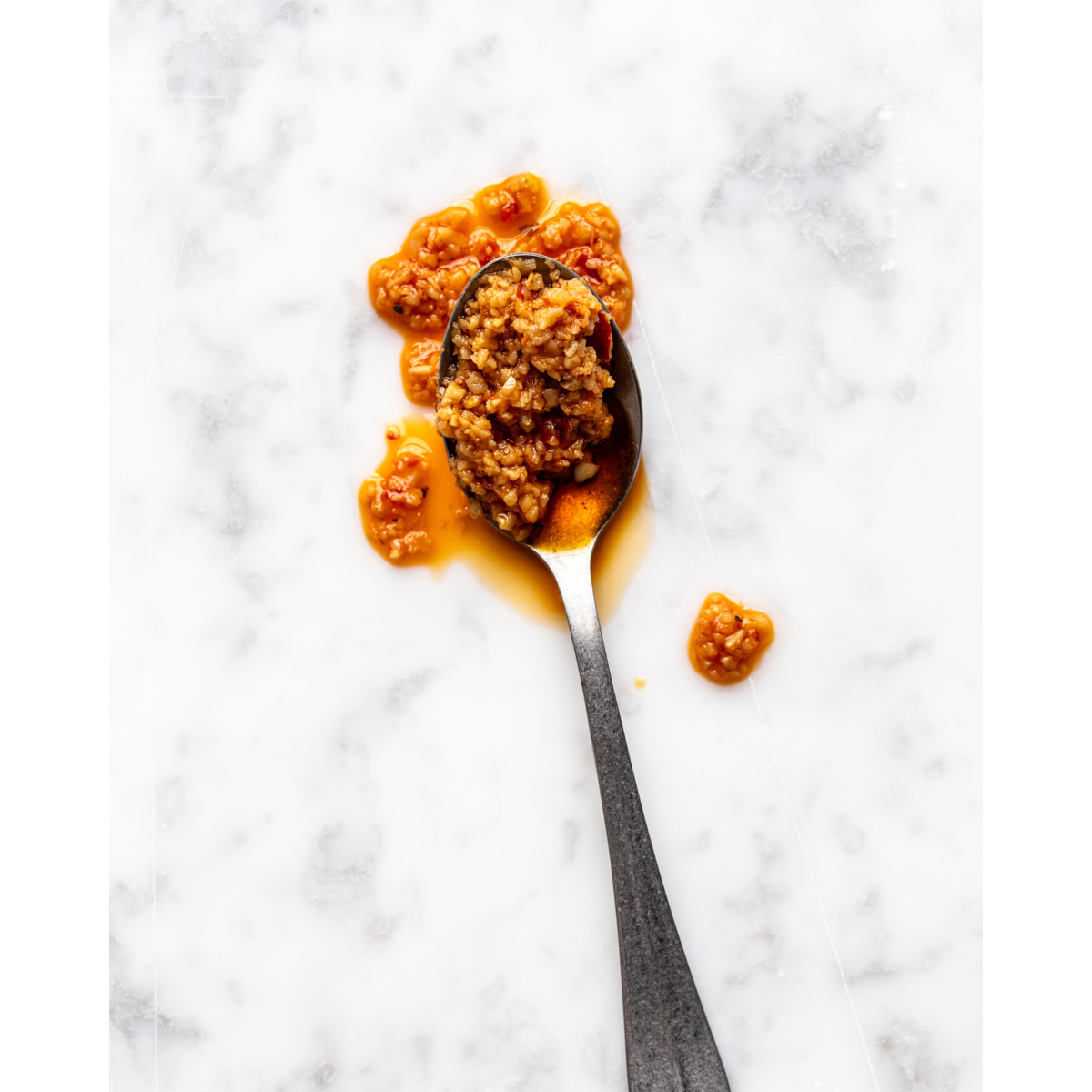 A metal spoon with Nicolas Vahé Spicy Garlic in Chilli Oil rests on a white marble surface, with some of the vibrant reddish-brown oil and condiment spilled around it.