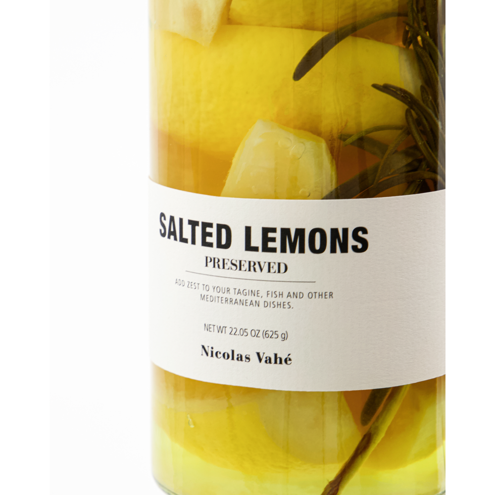 A close-up of a jar of Nicolas Vahé Salted Lemons, Preserved, displays yellow lemon pieces and herbs in liquid. The label recommends using them for tagine, fish, and Mediterranean dishes.