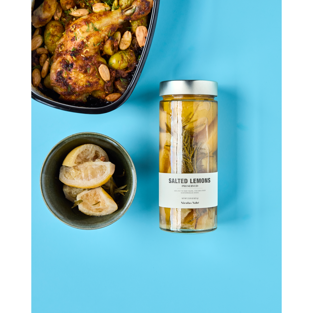 A jar of Nicolas Vahé Salted Lemons, Preserved, rests on a blue surface next to lemon wedges and roasted chicken with almonds and herbs—perfect for adding vibrant Mediterranean flavor to your dishes.