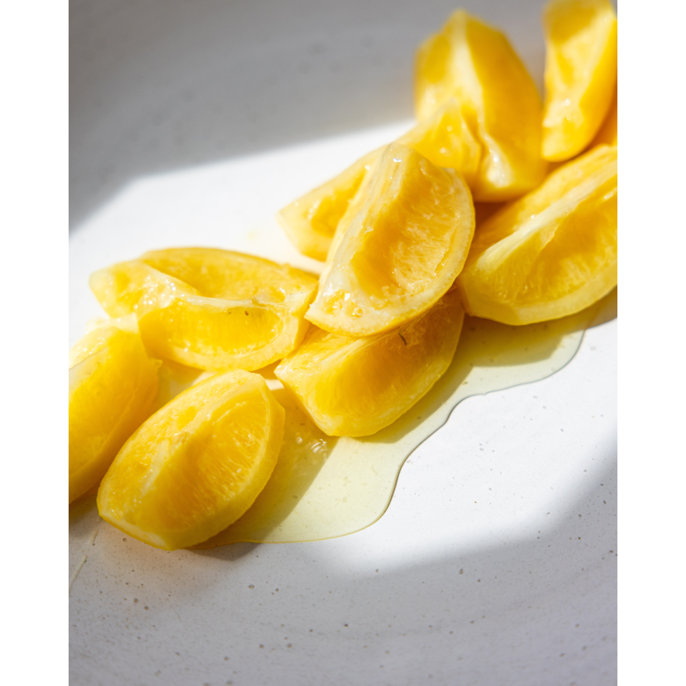 Wedges of Nicolas Vahé Salted Lemons, Preserved, are arranged on a white surface with their vibrant yellow hue and glistening juice pooling in the sunlight.