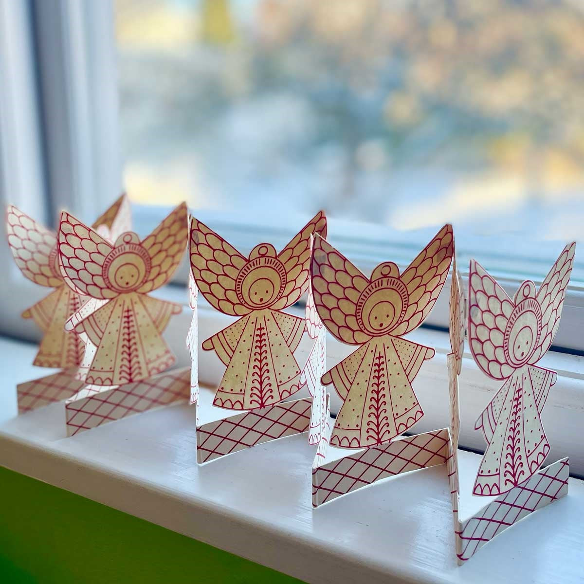 Four Folklore Angel Paper Foldout Christmas Decorations by A World of Craft, each featuring red Scandinavian-inspired patterns, stand on a windowsill against a softly blurred outdoor background—creating a handmade holiday display that captures the charm of Nordic tradition.