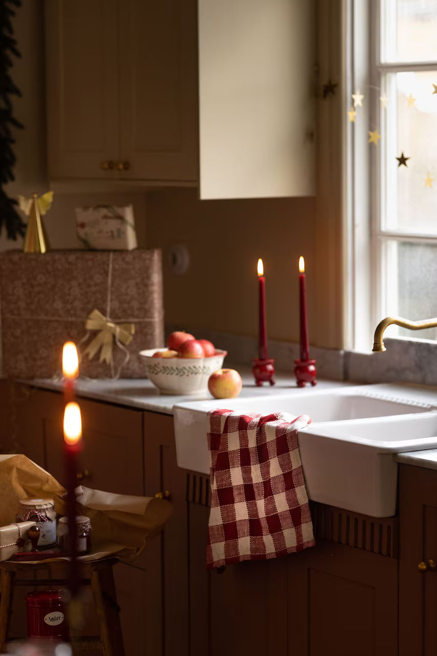 A cozy kitchen features a Strömshaga Ida Kitchen Towel in red and white check, draped over the sink, complemented by red candles, a bowl of apples, a wrapped gift, and warm light streaming in from the window for a festive and inviting atmosphere.