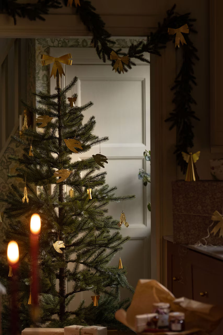 A sparsely decorated Christmas tree with gold ornaments is topped with the Strömshaga Tree Topper Bow in Brass, standing near a doorway. Two lit candles and wrapped gifts are visible in the foreground, while festive holiday decor—including garlands—are draped along the walls.