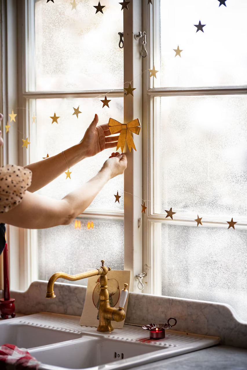 A person decorates a kitchen window with Strömshaga’s Ribbon Hanging Ornament in brass, creating a festive touch above the sink and complementing the brass faucet with elegant, cascading ribbon nearby.