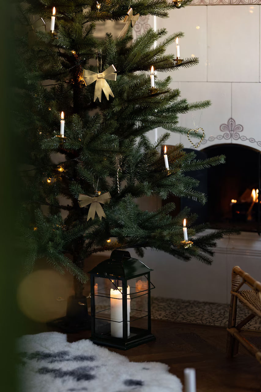 A Christmas tree decorated with candles, gold paper ornaments, and the elegant Ribbon Hanging Ornament in Brass by Strömshaga stands near a lit fireplace. A large white candle sits in a black lantern on the floor beside a cozy stool, while strands of decorative ribbon add an elegant touch to the festive scene.