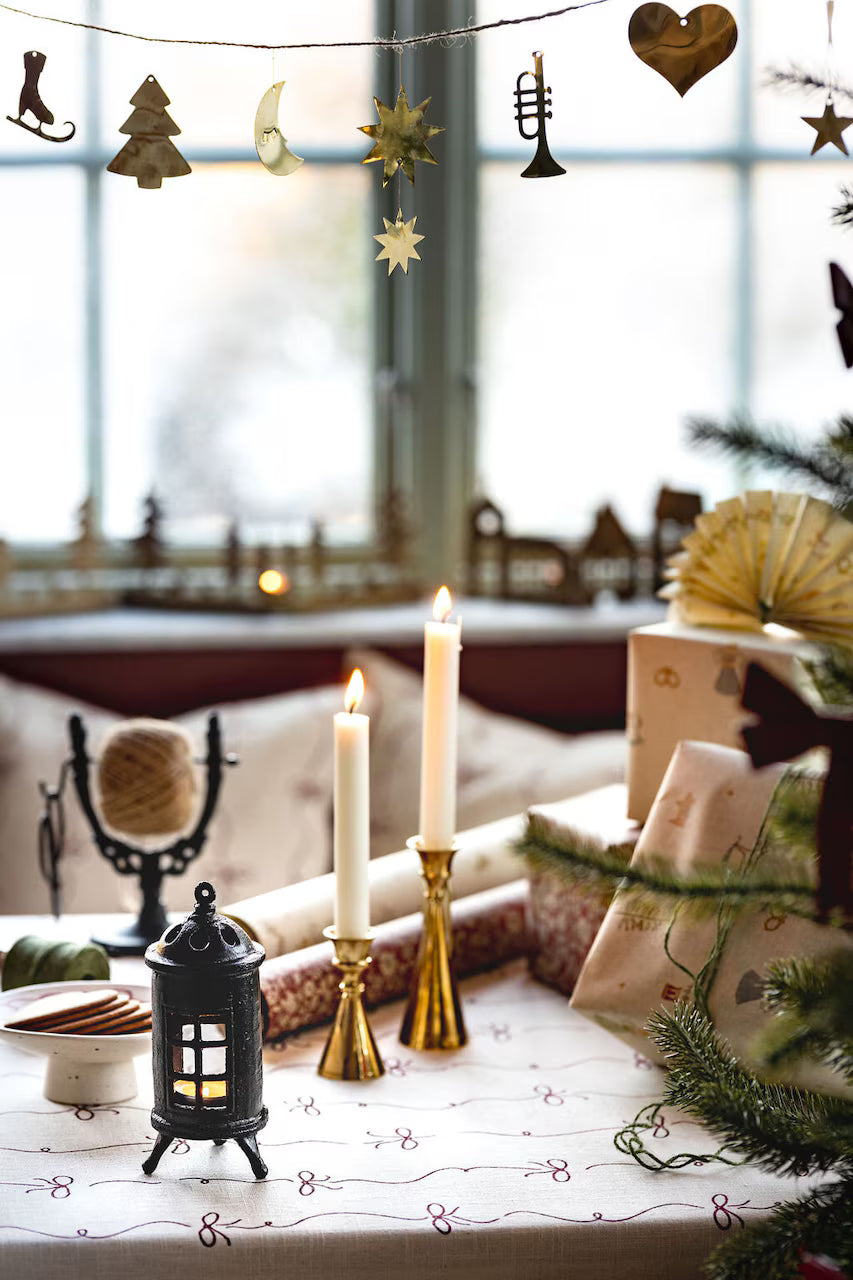 A festive table features two tea lights in the Strömshaga Stove Candleholder (cast iron), wrapped gifts, a lantern, and holiday décor by a window adorned with ornaments.