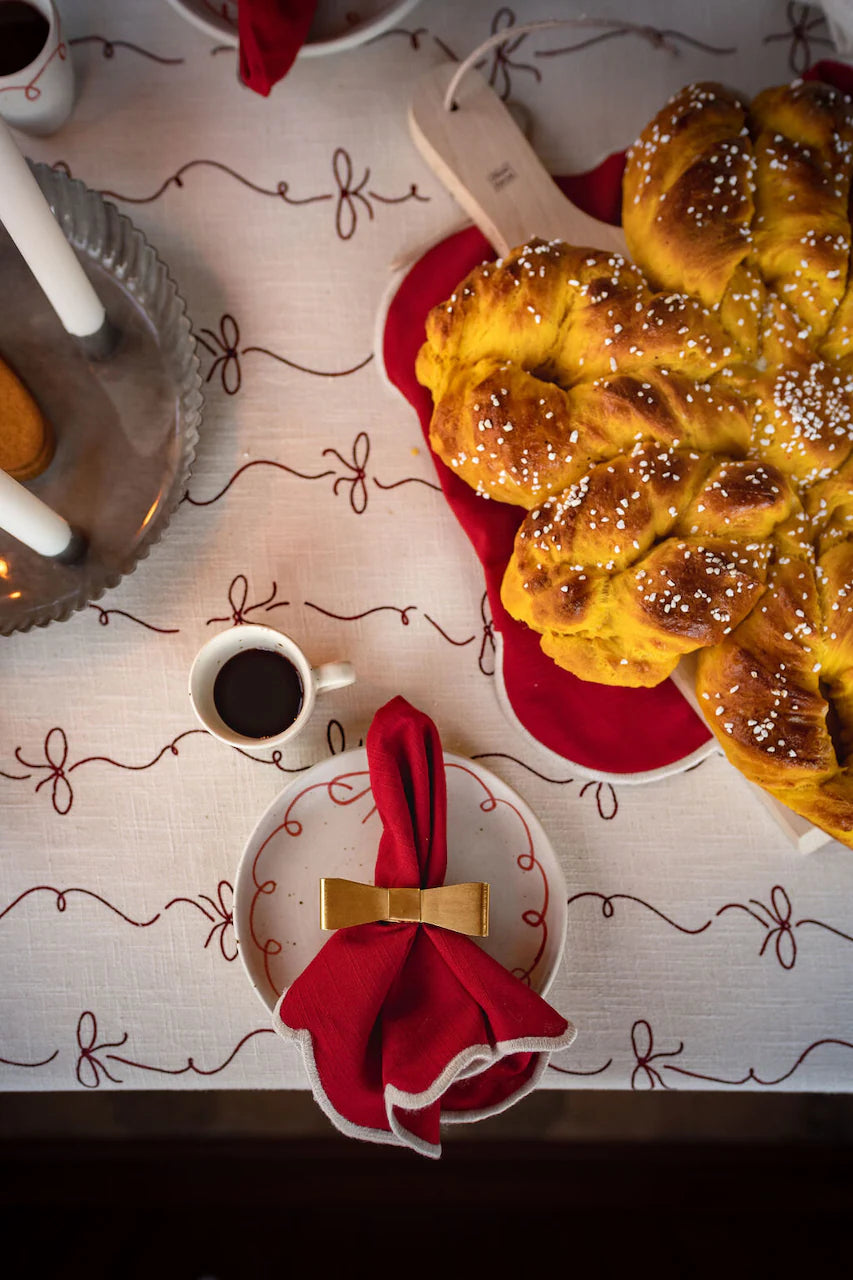 A braided sweet bread topped with pearl sugar, a cup of coffee, and a plate with a red napkin tied with a gold ribbon and finished with the Napkin Ring Bow Brass by Strömshaga are arranged on a festive white tablecloth.