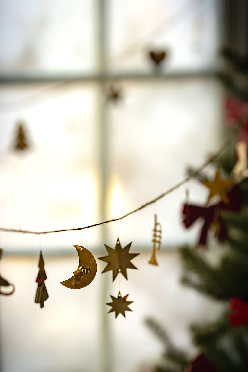 The Strömshaga Garland with Ornaments in Brass (2.7m) features classic Christmas motifs—crescent moon, stars, tree, and trumpet—hanging in front of a window with a glimpse of a decorated tree on the right.