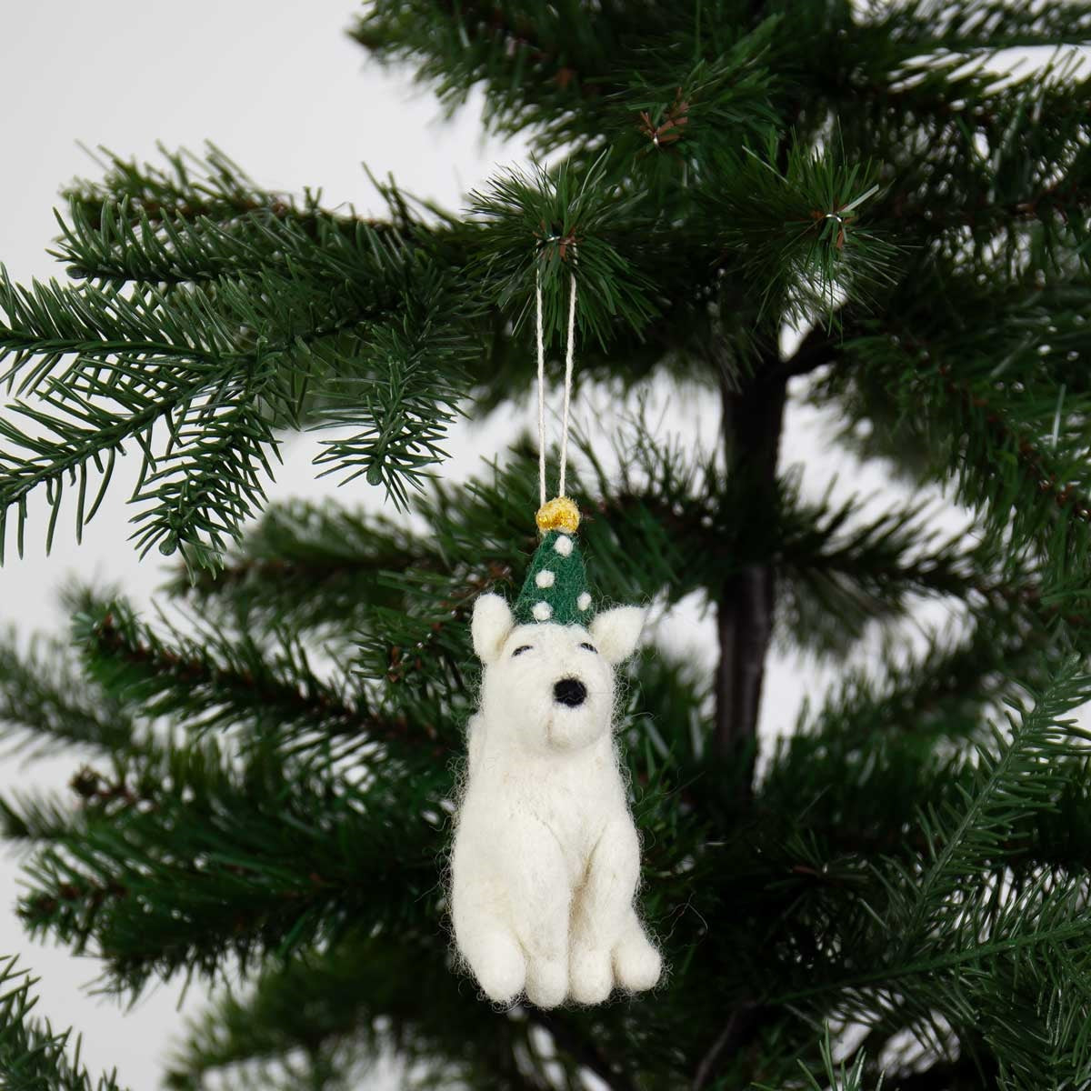 A handcrafted Party Dog Christmas Ornament, Handmade in Wool, white with a green polka-dot party hat, hangs from a festive Christmas tree branch.