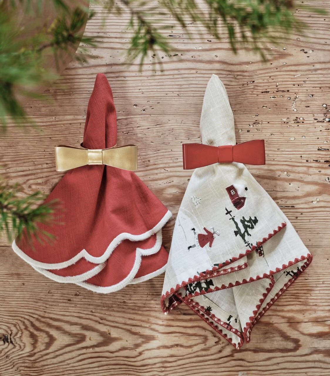Two napkins—one red with a Strömshaga Napkin Ring Bow Brass and one white with red trim and a Strömshaga Napkin Ring Bow Brass—are folded into dress shapes and placed on a wooden surface with pine branches partially visible, creating an elegant table setting.