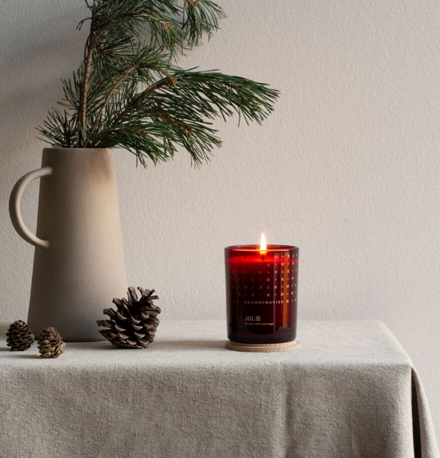 The Skandinavisk Seasonal Jul Scented Candle 50h, 200g sits on a cloth-covered table beside a ceramic vase with pine branches and three pinecones, capturing the cozy charm of a Scandinavian Christmas.