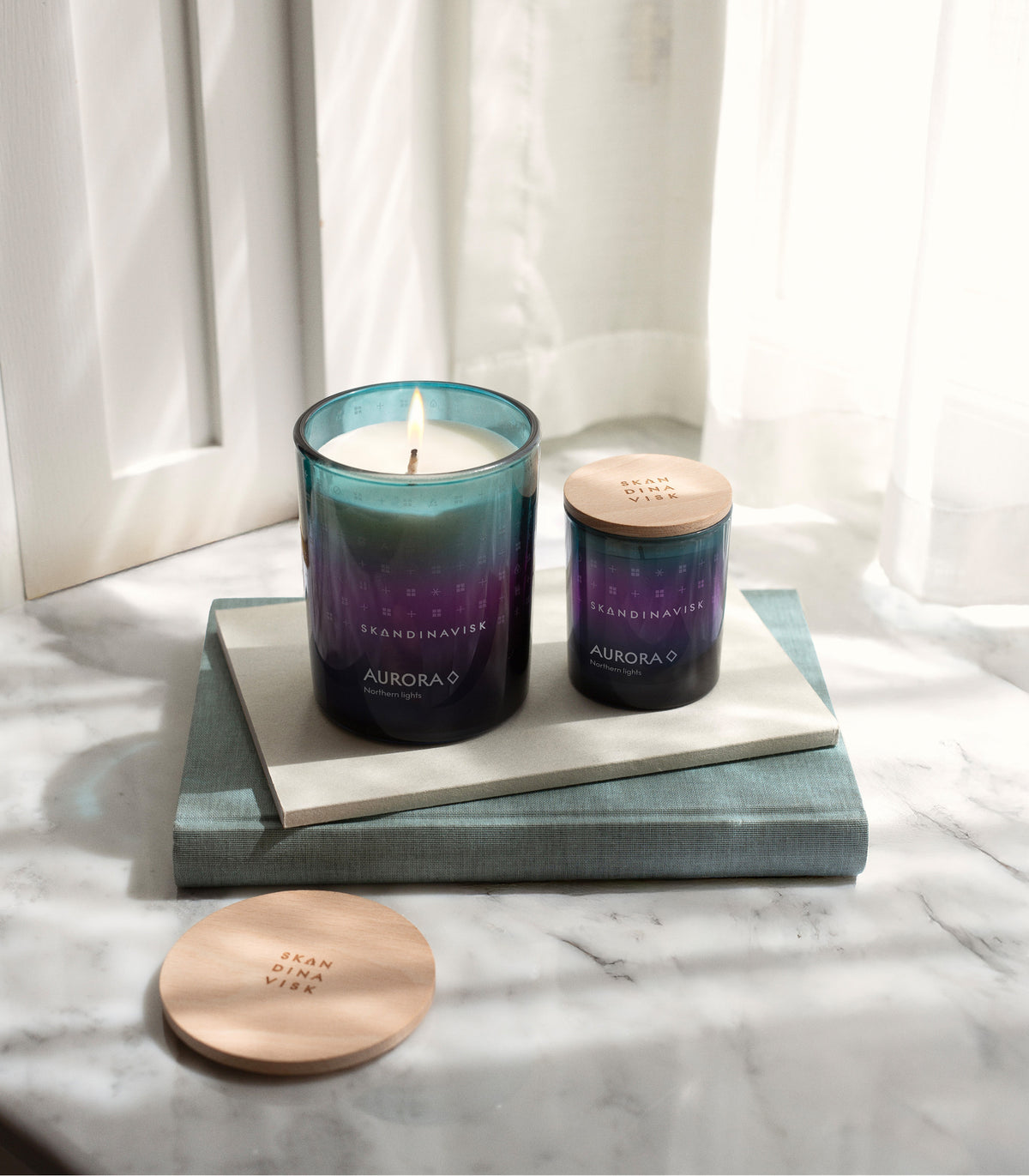 Two gradient glass Skandinavisk Seasonal Scented Candle Auroras (200 g), one glowing and one unlit, crafted from Swedish rapeseed wax with evocative notes of sandalwood and juniper berries, rest atop stacked books on a marble surface bathed in sunlight through sheer curtains. Nearby lie wooden lids—one engraved with Skandinavisk—capturing the elegant beauty inspired by the aurora borealis.