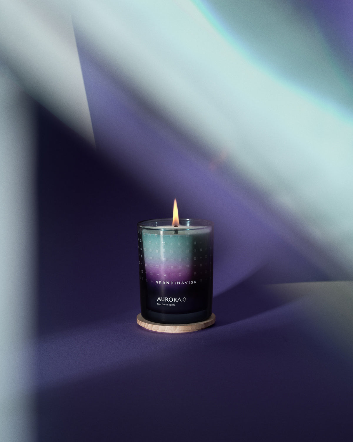 A lit Skandinavisk Seasonal Scented Candle Aurora, 200 g, rests on a wooden coaster, its gentle Swedish rapeseed wax delicately scented with sandalwood and juniper berries. Surrounded by soft purple lighting and blurred shapes, it creates a calm and cozy atmosphere.