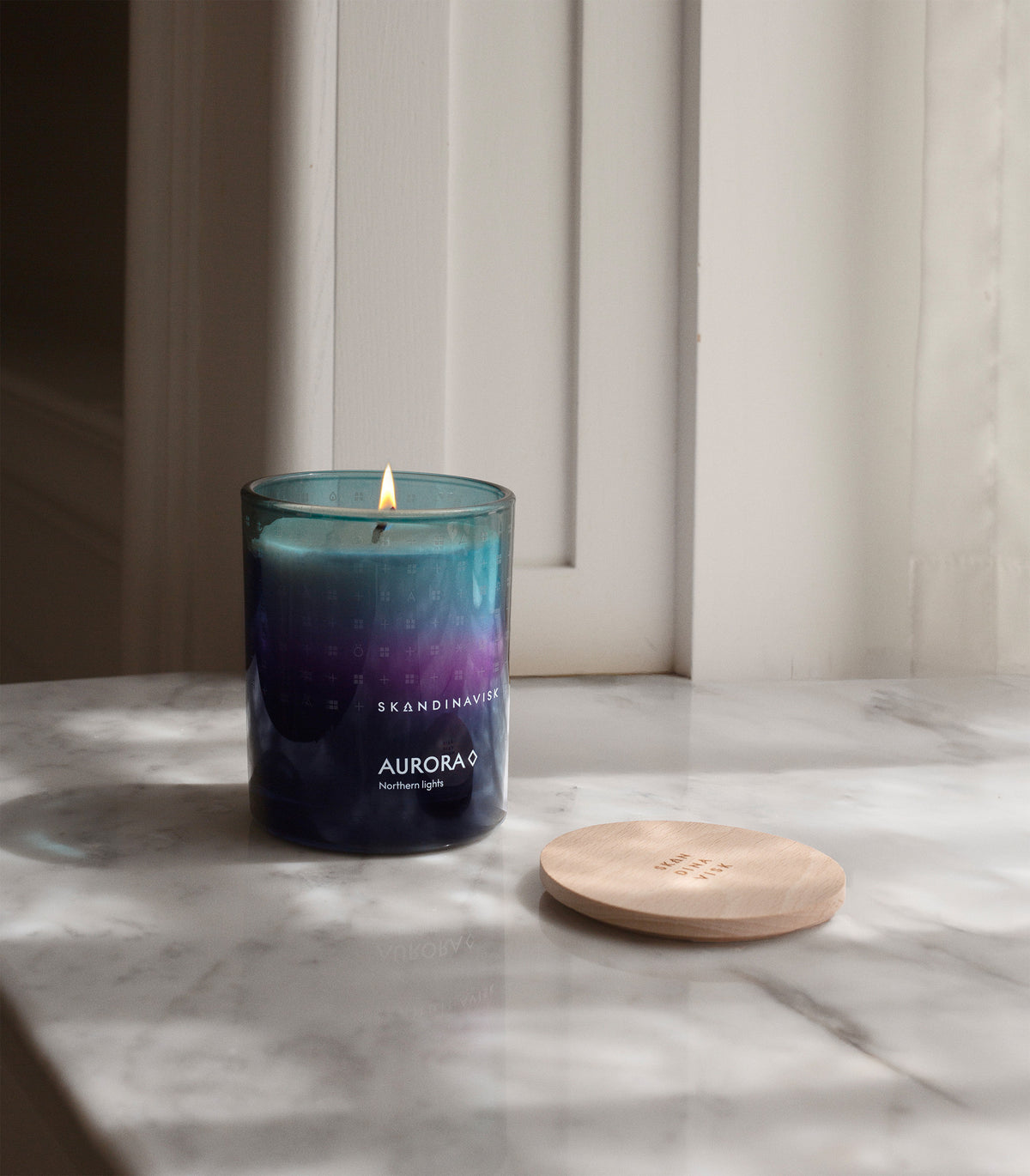 A lit Skandinavisk Seasonal Scented Candle Aurora, 200 g, made from Swedish rapeseed wax and fragranced with sandalwood and juniper berries, glows in its blue glass jar on a marble surface beside its wooden lid. Soft natural light filters through the window, enhancing the tranquil atmosphere.