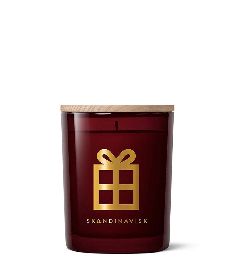 The Skandinavisk Seasonal Scented Candle Jul Gold Symbol (200 g) is presented in a burgundy glass jar with a wooden lid, adorned with a gold gift box icon and the brand name SKANDINAVISK displayed beneath it. Made from Swedish rapeseed wax, this Scandinavian Christmas candle fills your space with the cozy scent of gingerbread, perfect for creating seasonal warmth.