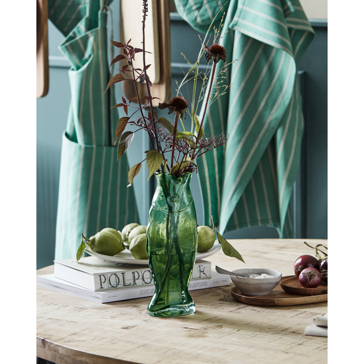 The House Doctor Vase Fish in Green Glass holds assorted flowers on stacked books beside green pears, a wooden tray of vegetables, and a green striped apron hanging in the background on a light wooden table.