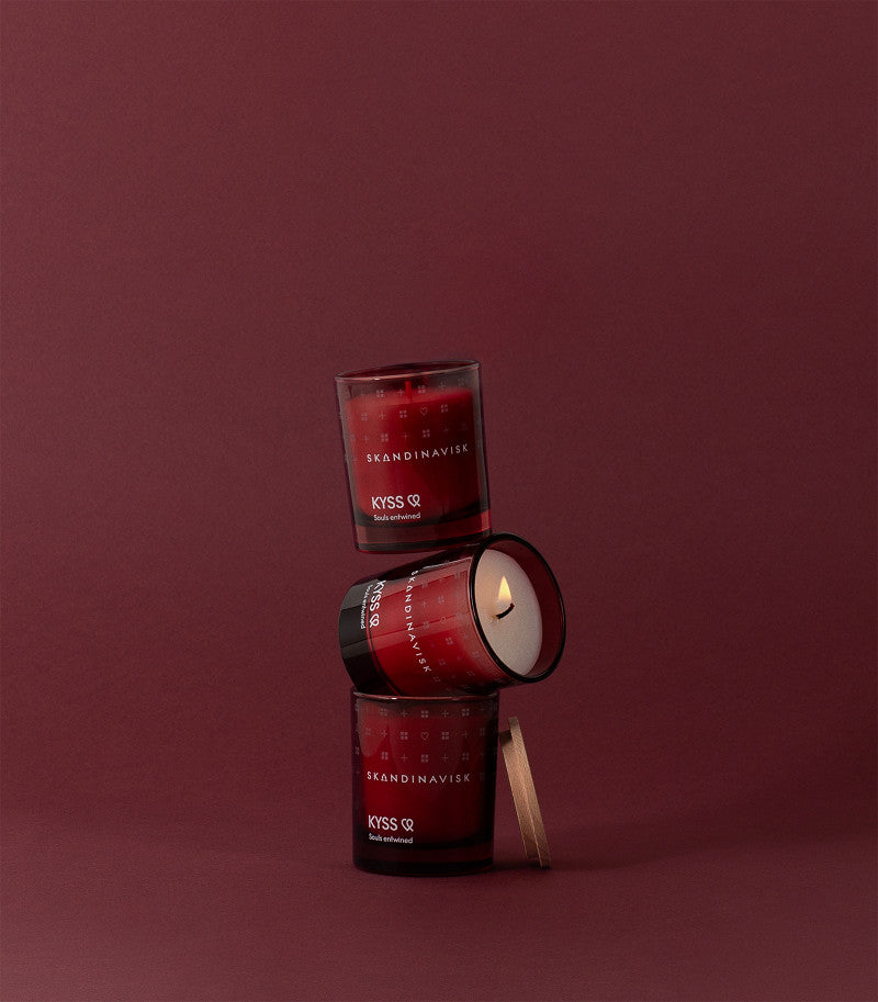 Three Skandinavisk Scented Candle Kyss 65g are stacked on a maroon background; the middle candle is lit and tilted diagonally, while the top and bottom candles remain upright.