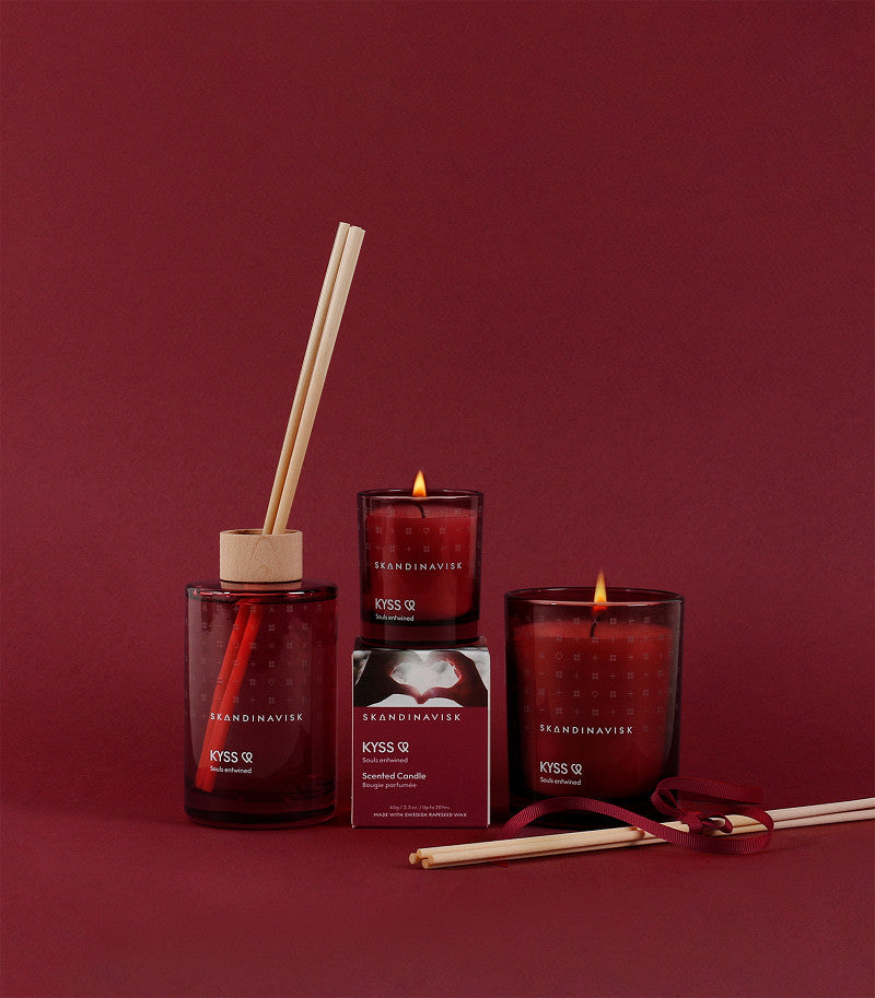 A Skandinavisk Scented Candle Kyss 65g, engraved beechwood lid, reed diffuser, and red box are arranged on a matching red background with reed sticks and ribbon.