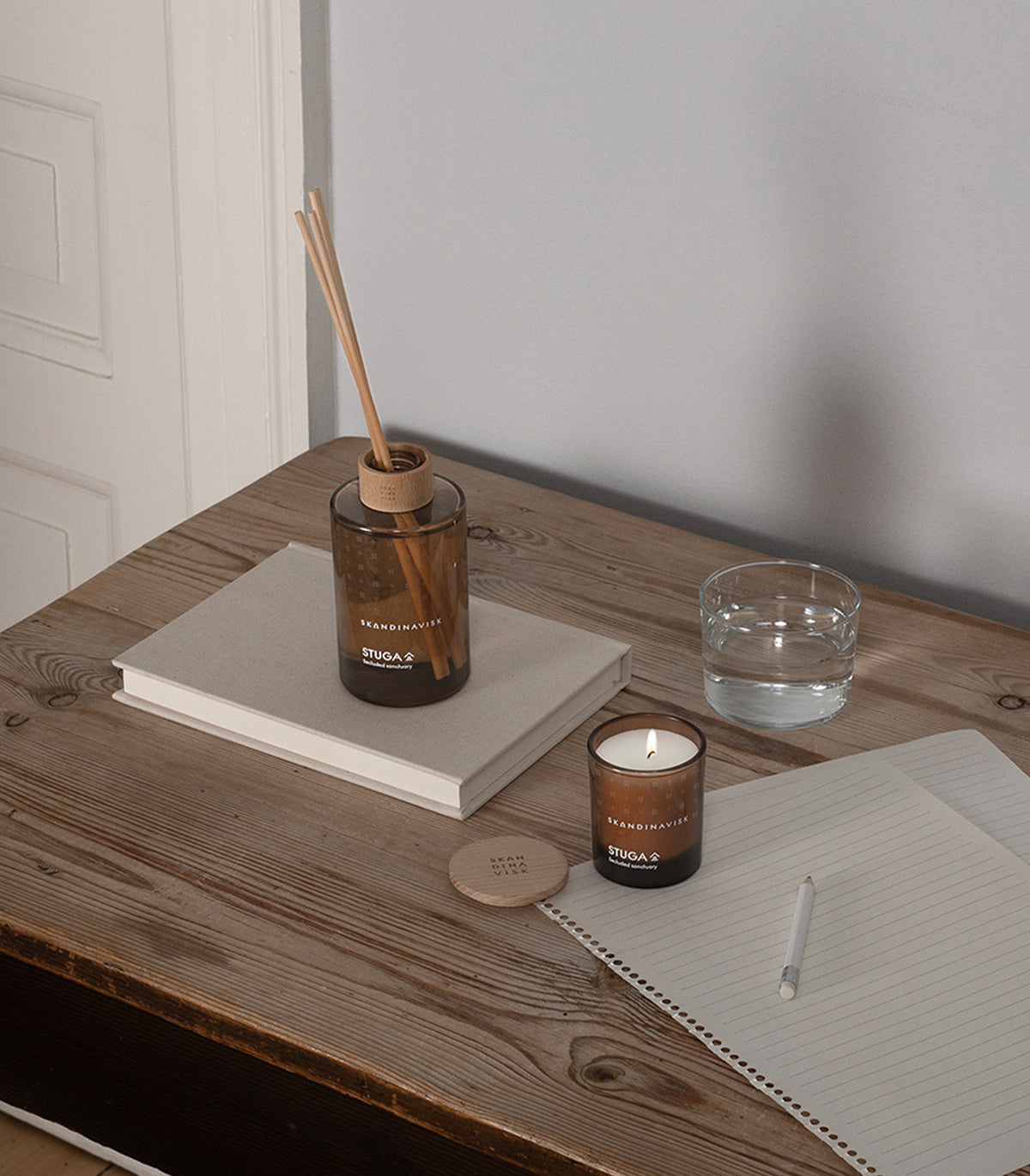 A minimal and calm setting featuring a wooden desk holds a notebook, pencil, closed book, glass of water, candle lid, scent diffuser emitting birch bark and woodland moss aromas, and the lit Skandinavisk Seasonal Scented Candle Stuga (65 g), crafted from Swedish rapeseed wax.