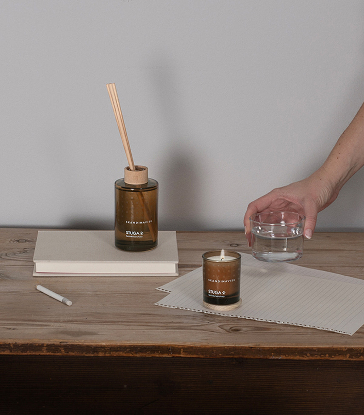 A hand holds a glass bowl of water above a wooden desk featuring the Skandinavisk Seasonal Scented Candle Stuga, 65 g, alongside a reed diffuser, two stacked notebooks, a pencil, and lined paper set against a plain gray wall.