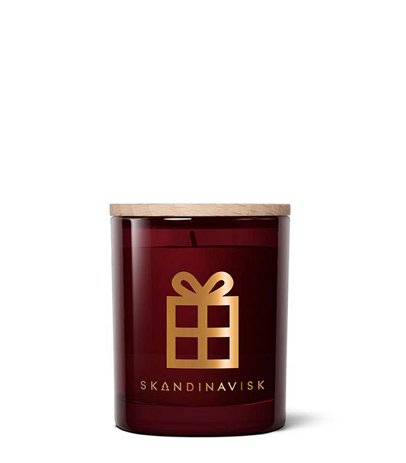 A 65 g Skandinavisk Seasonal Scented Candle Jul Gold Symbol, made from Swedish rapeseed wax, sits unlit against a white background. The burgundy glass jar features a wooden lid, a gold gift box symbol, and the Skandinavisk logo on the front for a festive Scandinavian Christmas touch.