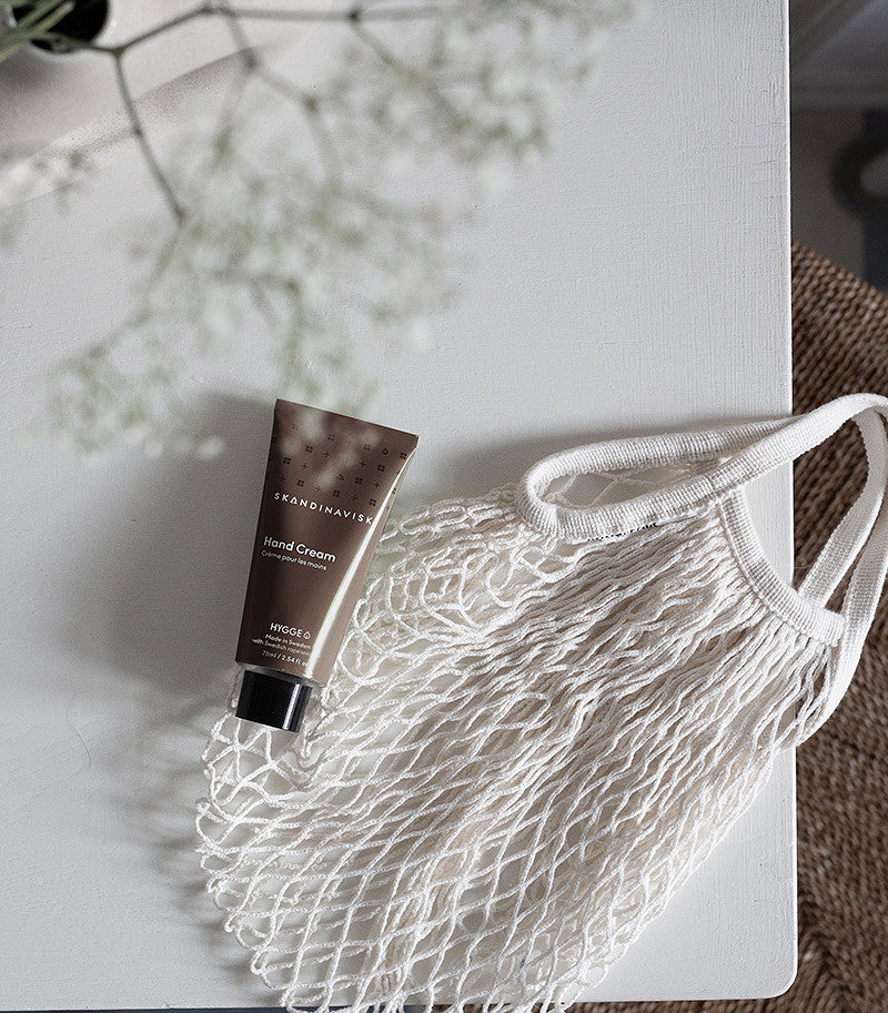 A tube of Skandinavisk Hand Cream Hygge (75 ml) lies on a white surface next to a white netted reusable bag, with a blurred plant in the top left corner.