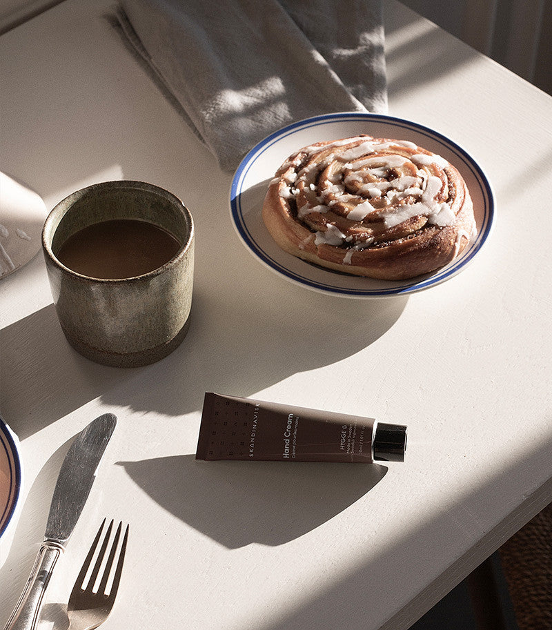 A cup of coffee, a cinnamon roll on a plate, the Skandinavisk Mini Hand Cream Hygge (30 ml), and cutlery are arranged on a white table in sunlight.