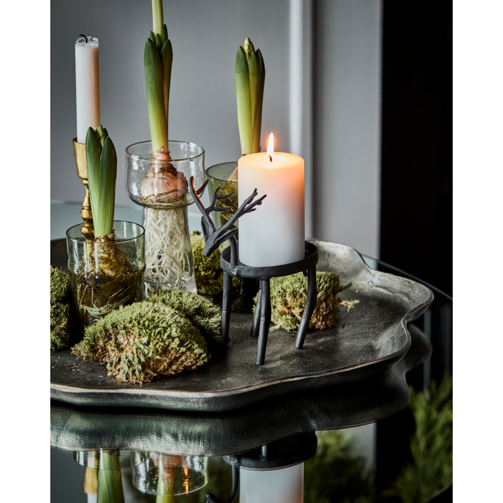 A black Tealight Holder Deer in Iron by House Doctor with a lit white pillar candle, displayed on a tray with potted plants and moss as part of a Christmas display.