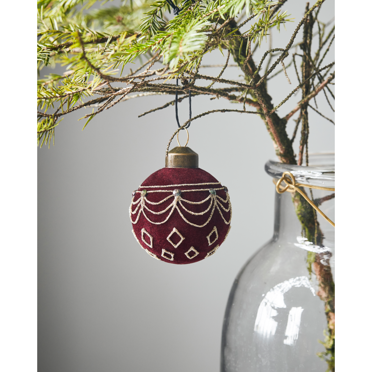 The House Doctor Christmas Ornament Bauble in Velour Red / Brown hangs from a sparse branch set in a clear glass vase, with a plain background.