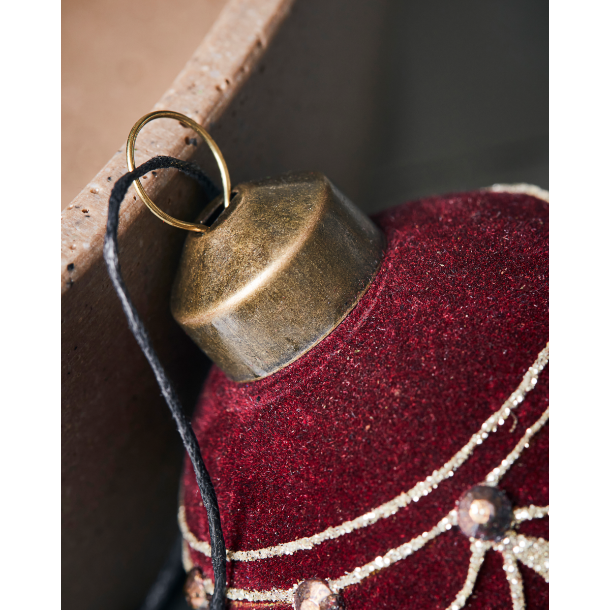 Close-up of the House Doctor Christmas Ornament Bauble, Velour Red / Brown, featuring gold detailing and a metal cap, hanging by a black string near a brown surface.