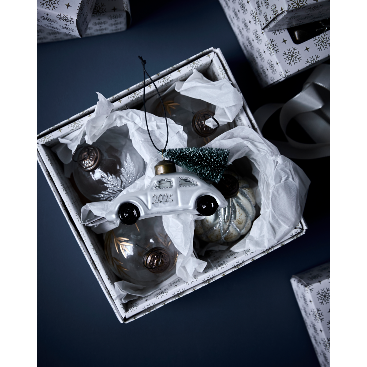 A House Doctor box holds mouth-blown glass ornaments, featuring the 2025 Ornament Car Bauble with Tree in white & silver, all nestled in soft white tissue paper.