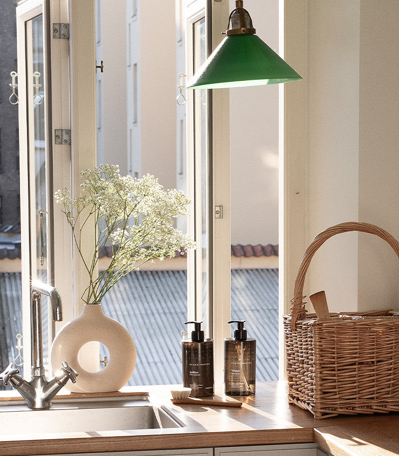 A sunlit kitchen windowsill features a white vase of flowers, a wicker basket, a green pendant lamp overhead, and two Skandinavisk Hand & Body Lotion Hygge (500 ml) dispensers, reflecting the serene essence of Scandinavian skincare.