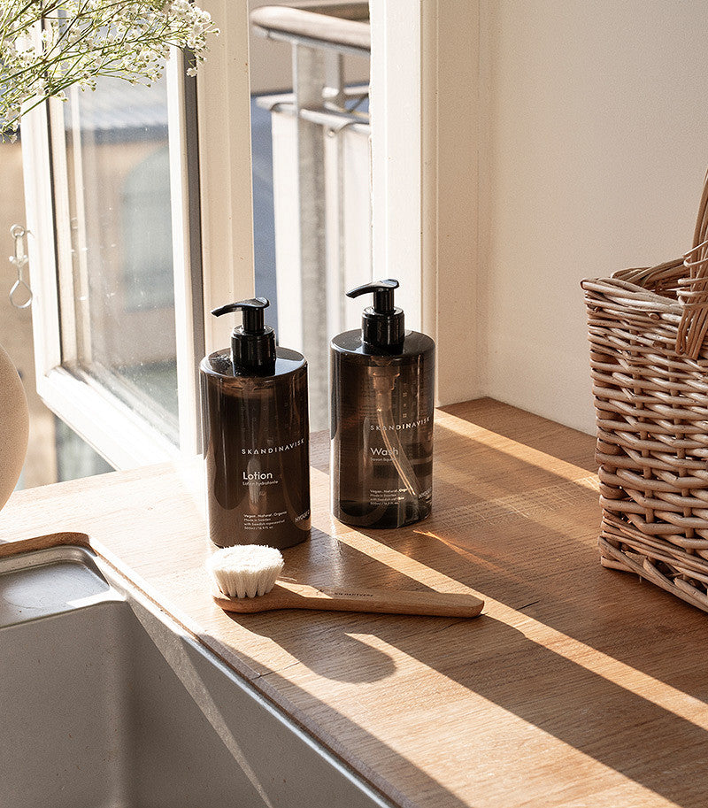 Two pump bottles labeled “Lotion” and “Wash”—including the Skandinavisk Hand & Body Lotion Hygge, 500 ml, enriched with organic aloe vera—rest on a sunwashed wooden counter near a window, accompanied by a wooden-handled brush and a wicker basket. Gentle natural light floods the scene, evoking the warm, inviting ambiance of Scandinavian skincare rituals.