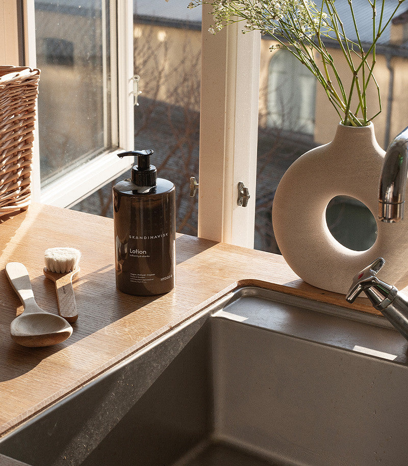 Sunlight streams through a kitchen window onto a wooden countertop, where a bottle of Skandinavisk Hand & Body Lotion Hygge (500 ml), enriched with organic aloe vera, sits among wooden utensils, a brush, a wicker basket, and a beige vase filled with greenery beside the stainless steel sink.