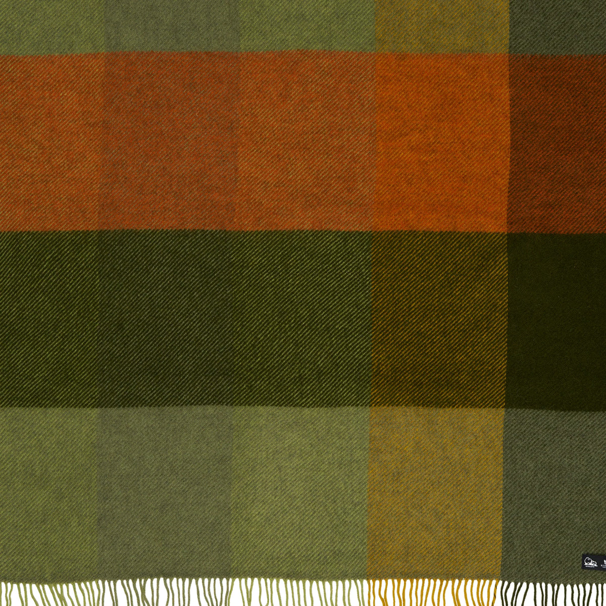 The Klippan Yllefabrik Gotland Brushed Gotland & Lambswool Throw in Check Green features a Swedish-designed plaid pattern in green, orange, brown, and yellow hues. The blanket is finished with a white fringe along the bottom edge and includes a small dark label in the lower right corner.