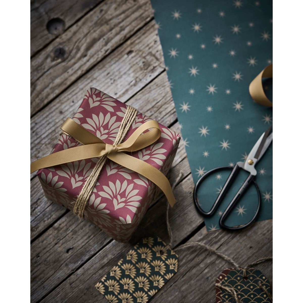A wrapped gift with gold ribbon sits on a wooden surface beside scissors, House Doctor gift tags, kraft paper, and double sided red & green wrapping paper with gold by House Doctor.