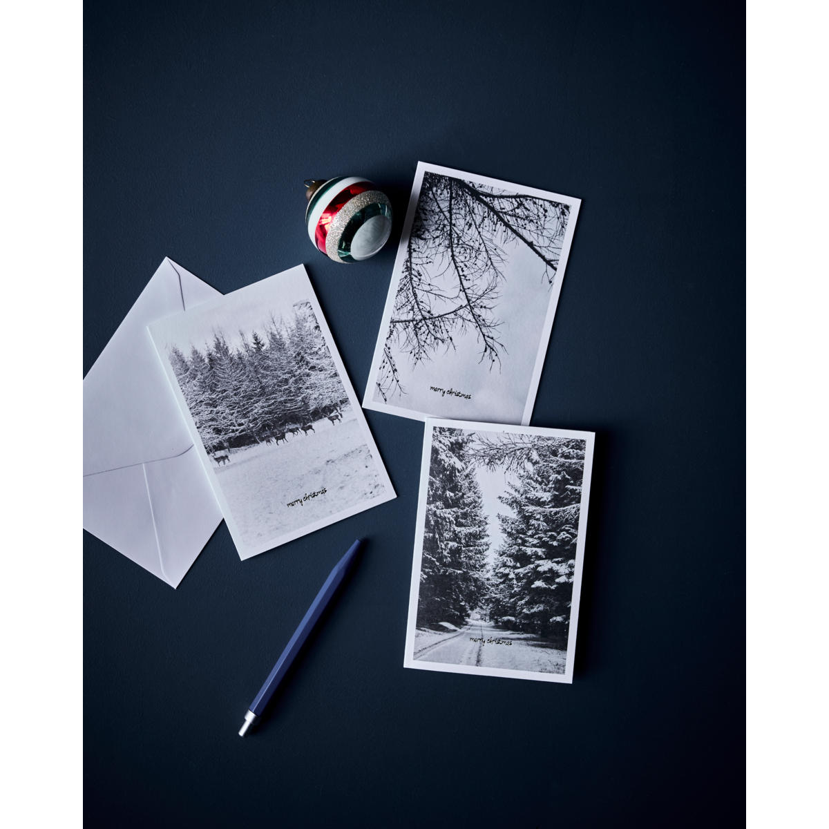 A set of six winter greeting cards by House Doctor, a blue pen, a white envelope, and a striped ornament are displayed on a dark surface.