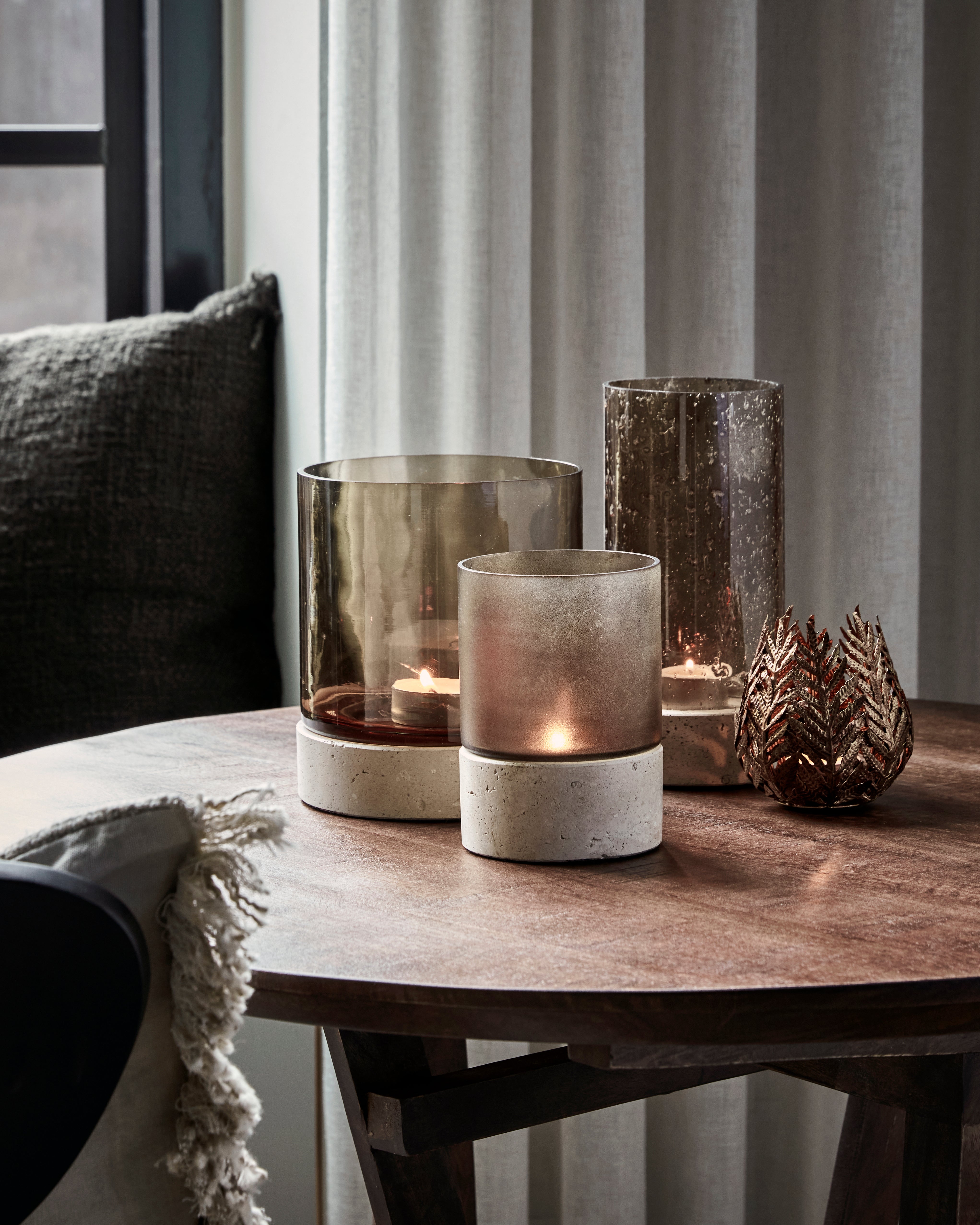 Three elegant Candle Holder Rivo w. Brown Glass by House Doctor, each handmade with mouthblown glass and sturdy travertine bases, feature a decorative metallic leaf ornament and are arranged on a round wooden table near a window with sheer curtains and a dark cushion in the background.
