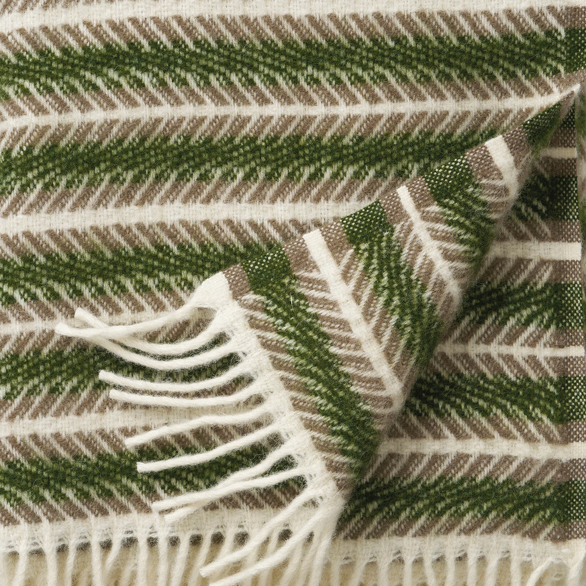 The Oscar Multi Green Lambswool Throw by Klippan Yllefabrik features green, brown, and white herringbone stripes with a white fringe. One corner is folded to display the underside of the pattern. Crafted from eco lamb’s wool, it offers exceptional softness and warmth.