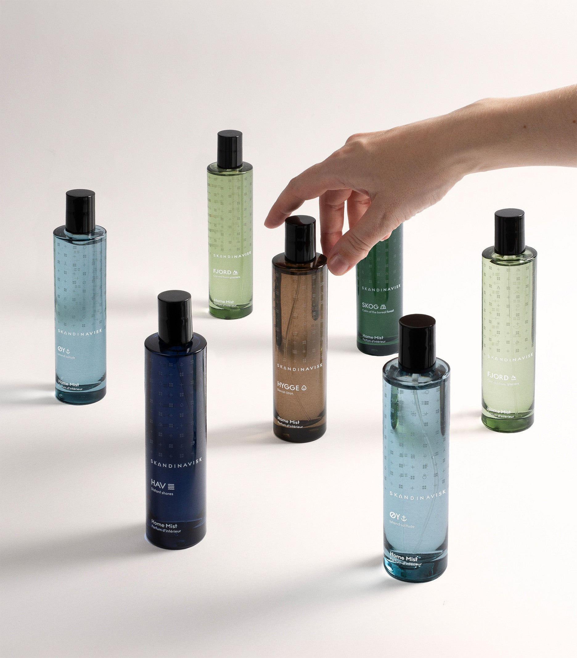 Seven bottles from Skandinavisk’s home fragrance collection are arranged on a white surface. A hand reaches for the brown bottle labeled “Home Mist Hygge, 100 ml,” capturing the comforting essence of Scandinavian hygge. The bottles display shades of blue, green, and beige, creating an inviting and tranquil atmosphere reminiscent of black tea perfume notes drifting through a peaceful home.