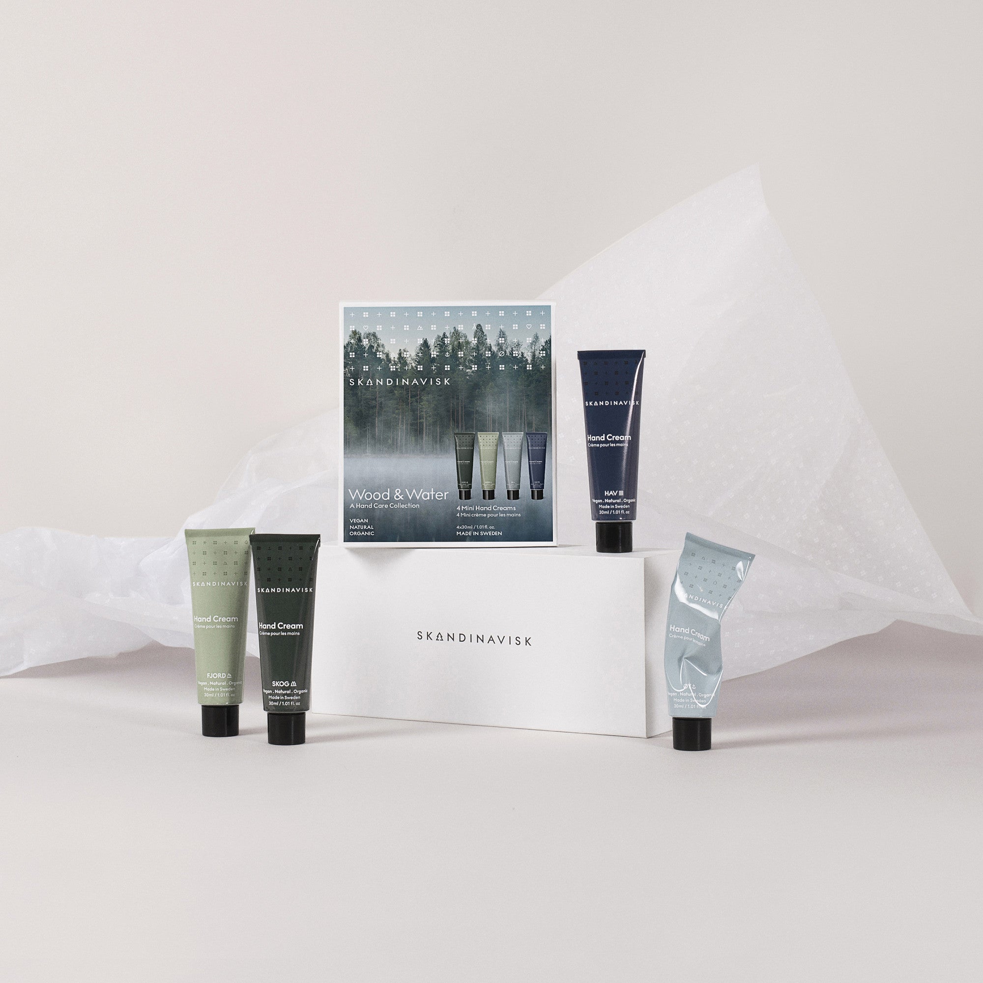 Skandinavisk’s “Giftset Hand Creams Wood & Water,” featuring four mini 30ml hand creams—Skog, Fjord, Oy, and Hav—is beautifully displayed on and around a branded white box amidst soft tissue paper and a neutral backdrop. These Scandinavian hand care essentials are thoughtfully crafted with natural ingredients to provide gentle nourishment.