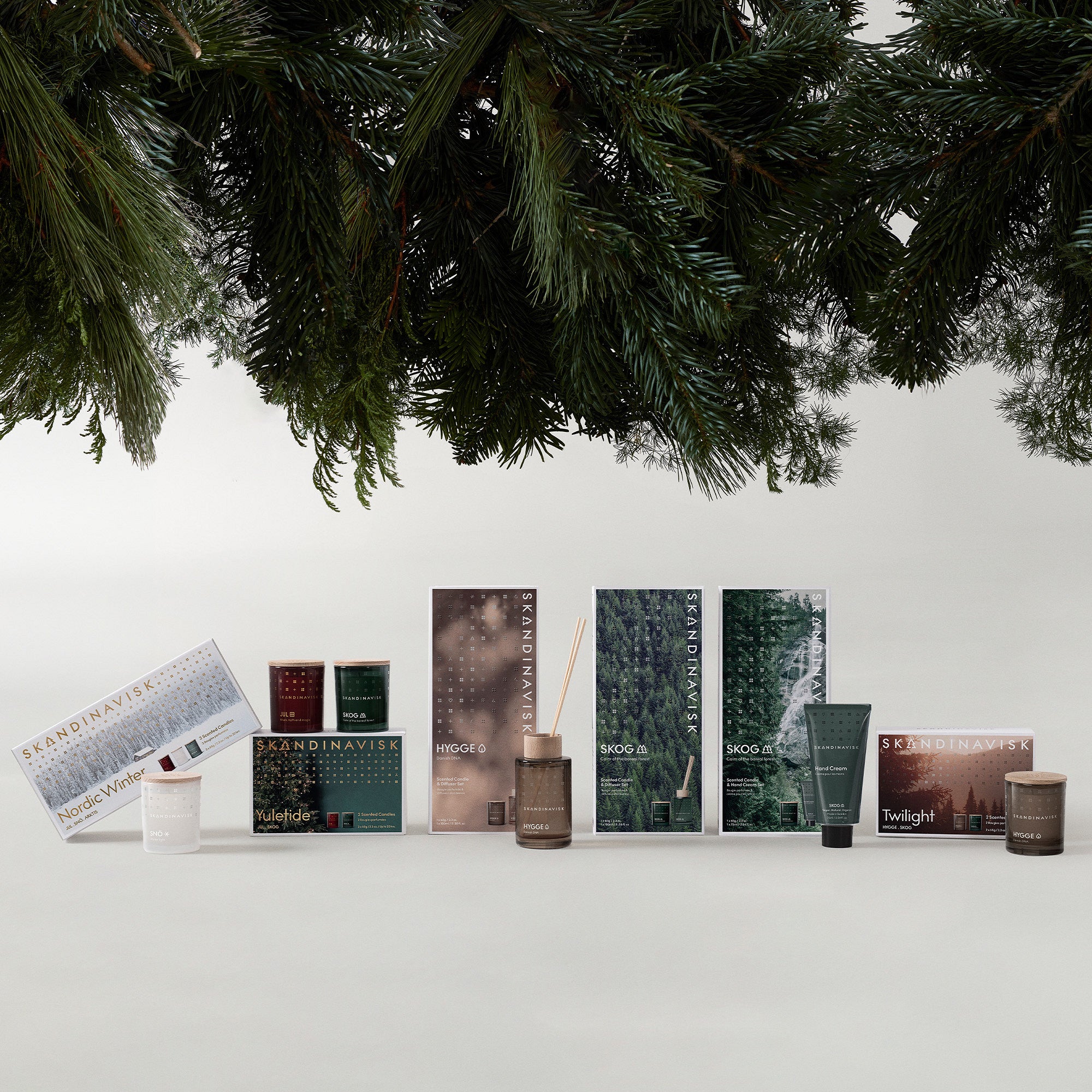 A display of Skandinavisk candles, diffusers, and packaging arranged beneath hanging pine branches creates a natural, forest-inspired theme. The centerpiece is the Giftset Scented Candle Twilight with 2 Mini Candles Skog & Hygge by Skandinavisk, evoking an inviting Nordic ambiance.