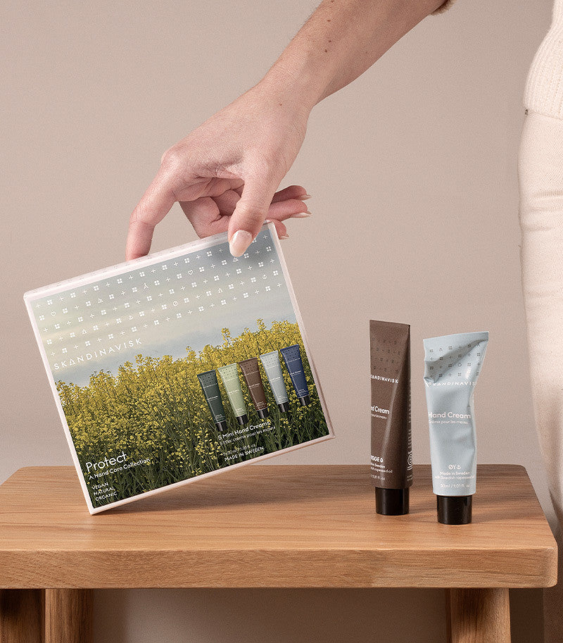 A hand holds the Skandinavisk Giftset Mini Hand Cream Protect, a set of five beautifully packaged tubes inspired by nature—SKOG, FJORD, ØY, HAV, and HYGGE. Two of the hand creams made with natural ingredients such as organic aloe vera stand upright on a wooden stool beside the person.