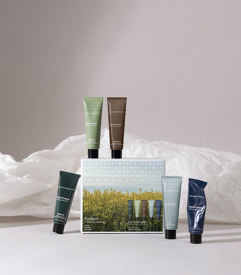 Five Skandinavisk Giftset Mini Hand Cream tubes—featuring the scents Skog, Fjord, Oy, Hav, and Hygge—are arranged around and atop a box adorned with yellow field flowers. Each vegan cream is crafted with natural ingredients such as organic aloe vera and delivers nourishing moisturisation for your hands. The display rests on a neutral background of crumpled white paper.