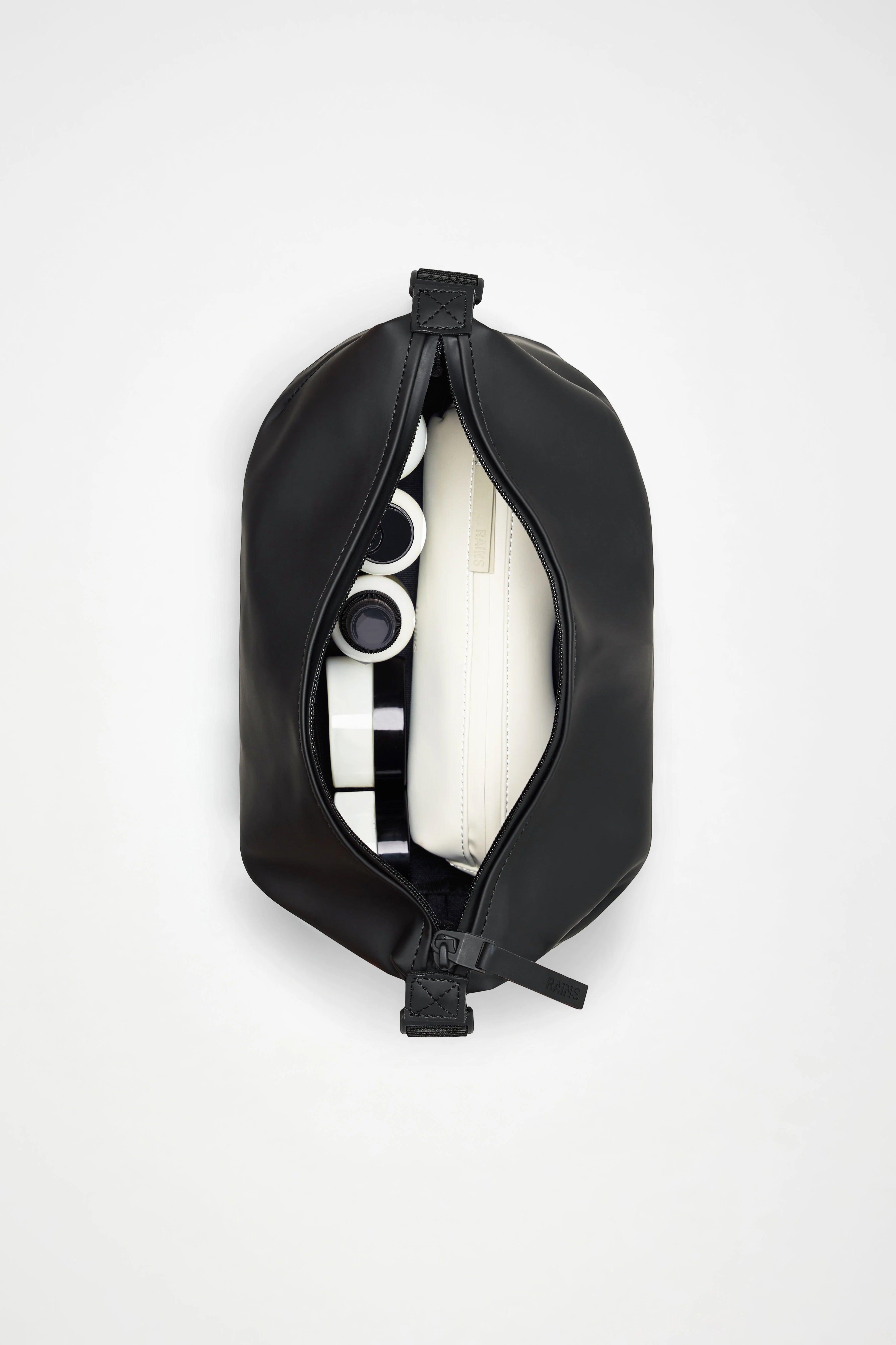 A green Rains Hilo Wash Bag W3 by Rains, partially unzipped, displays neatly organized contents inside—including a white rectangular case and black-and-white cylindrical items—all arranged against a clean white backdrop.