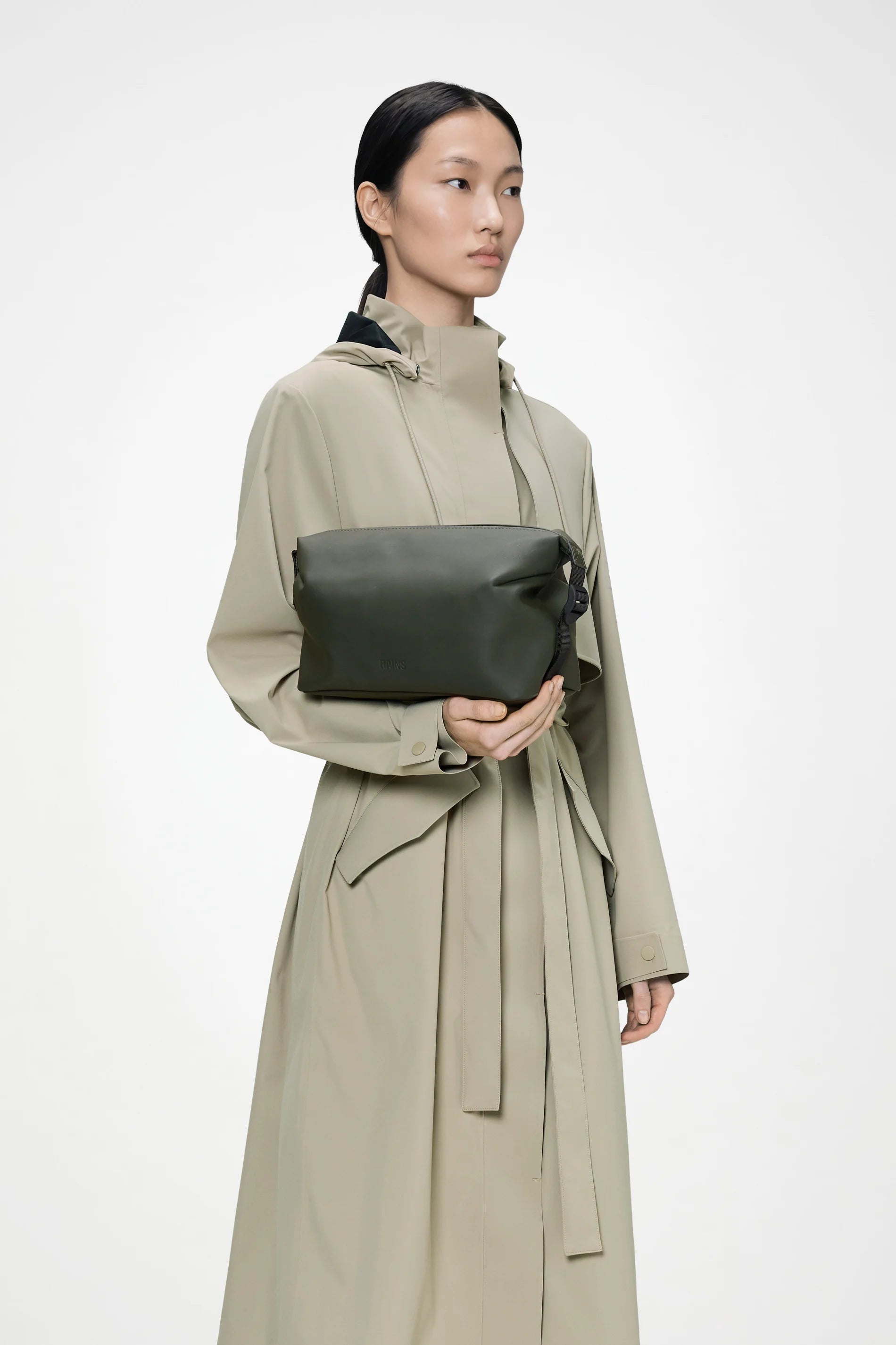 A person wearing a light beige trench coat holds the Rains Hilo Wash Bag W3 in Green by Rains across their chest, standing against a plain white background.