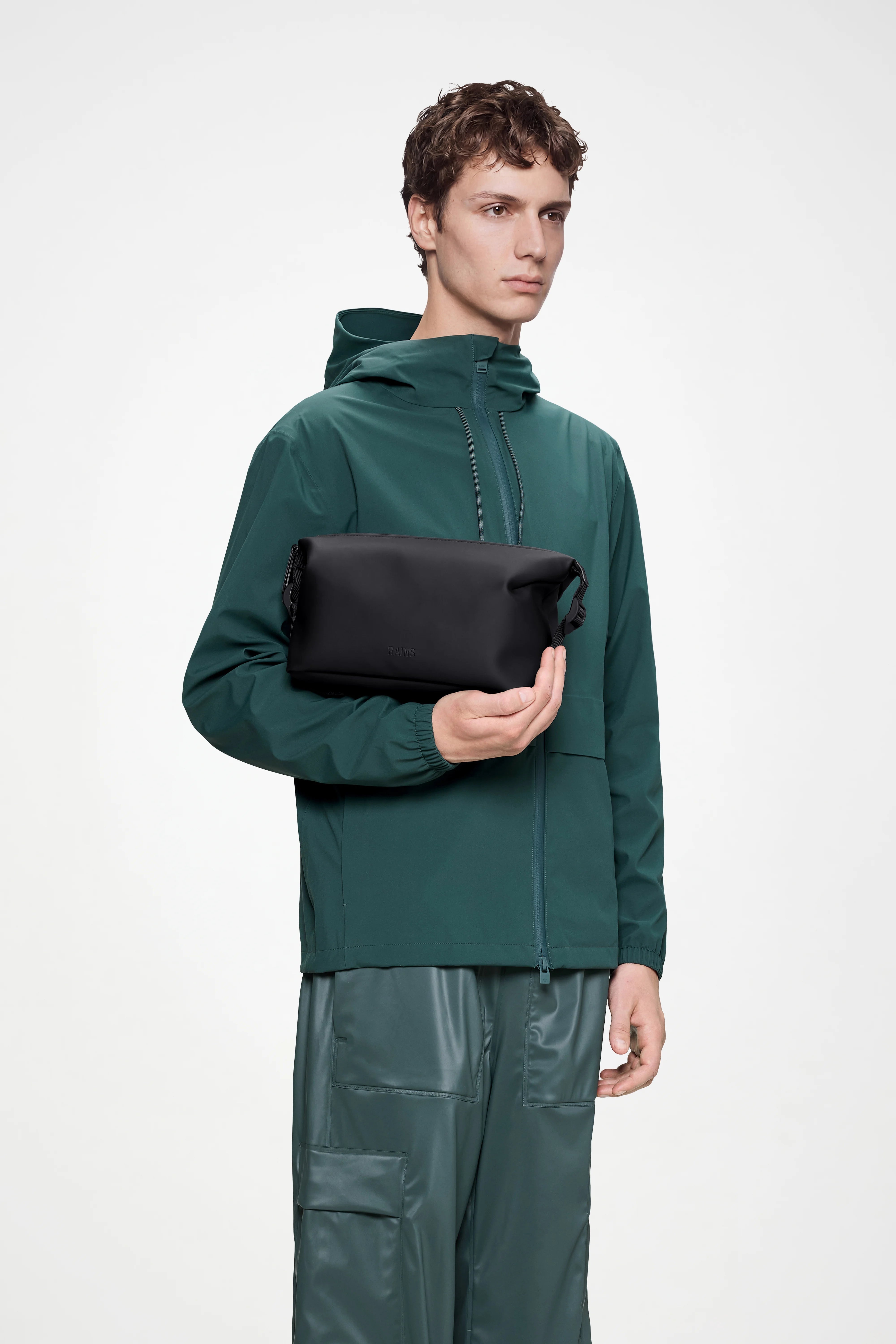 A person dressed in a green hooded jacket and matching pants holds the Rains Hilo Wash Bag W3 in Black by Rains against a plain white background.