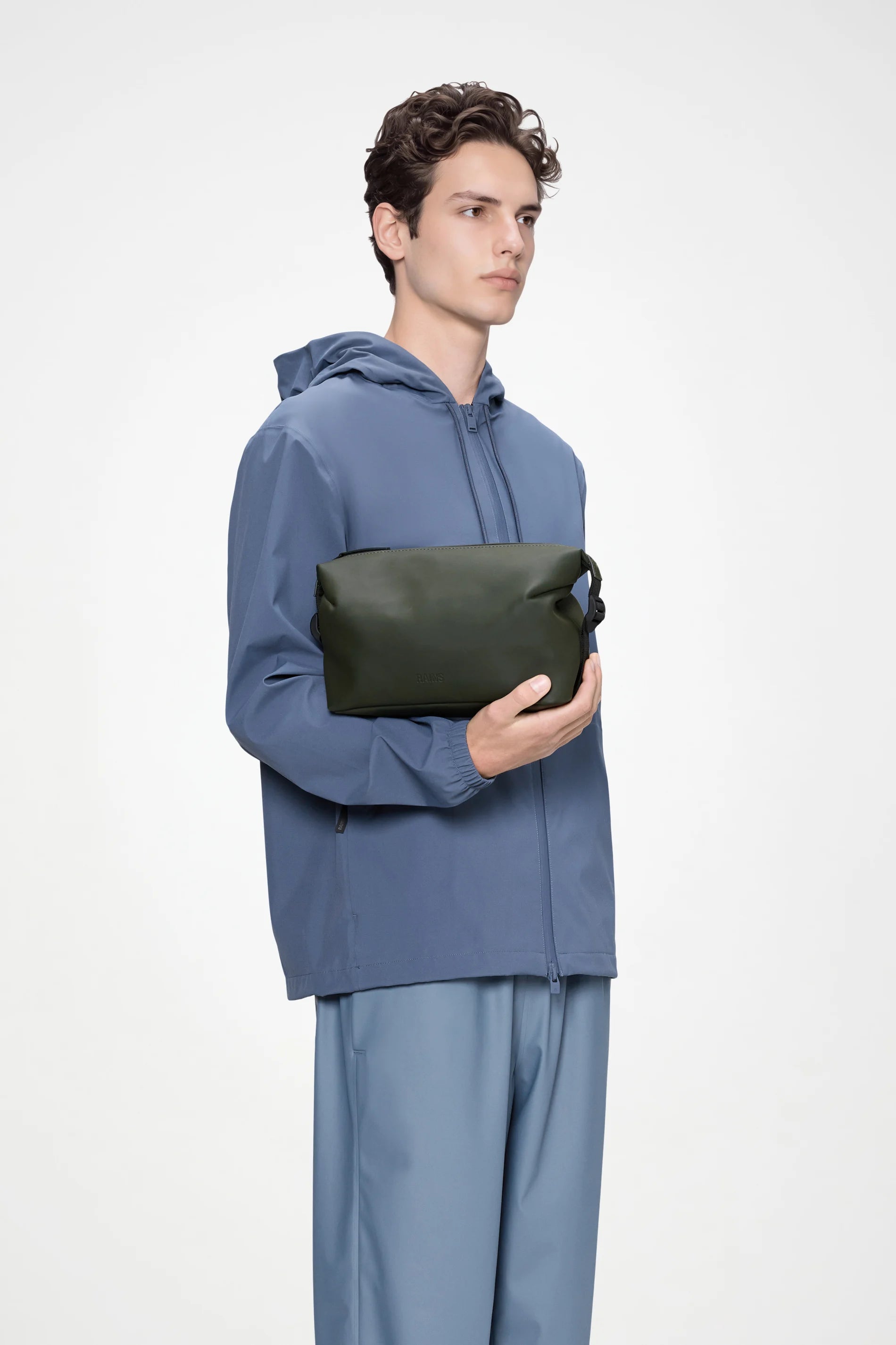A person in a blue hooded jacket and matching pants holds the Rains Hilo Wash Bag W3 in Green by Rains against a plain light background.