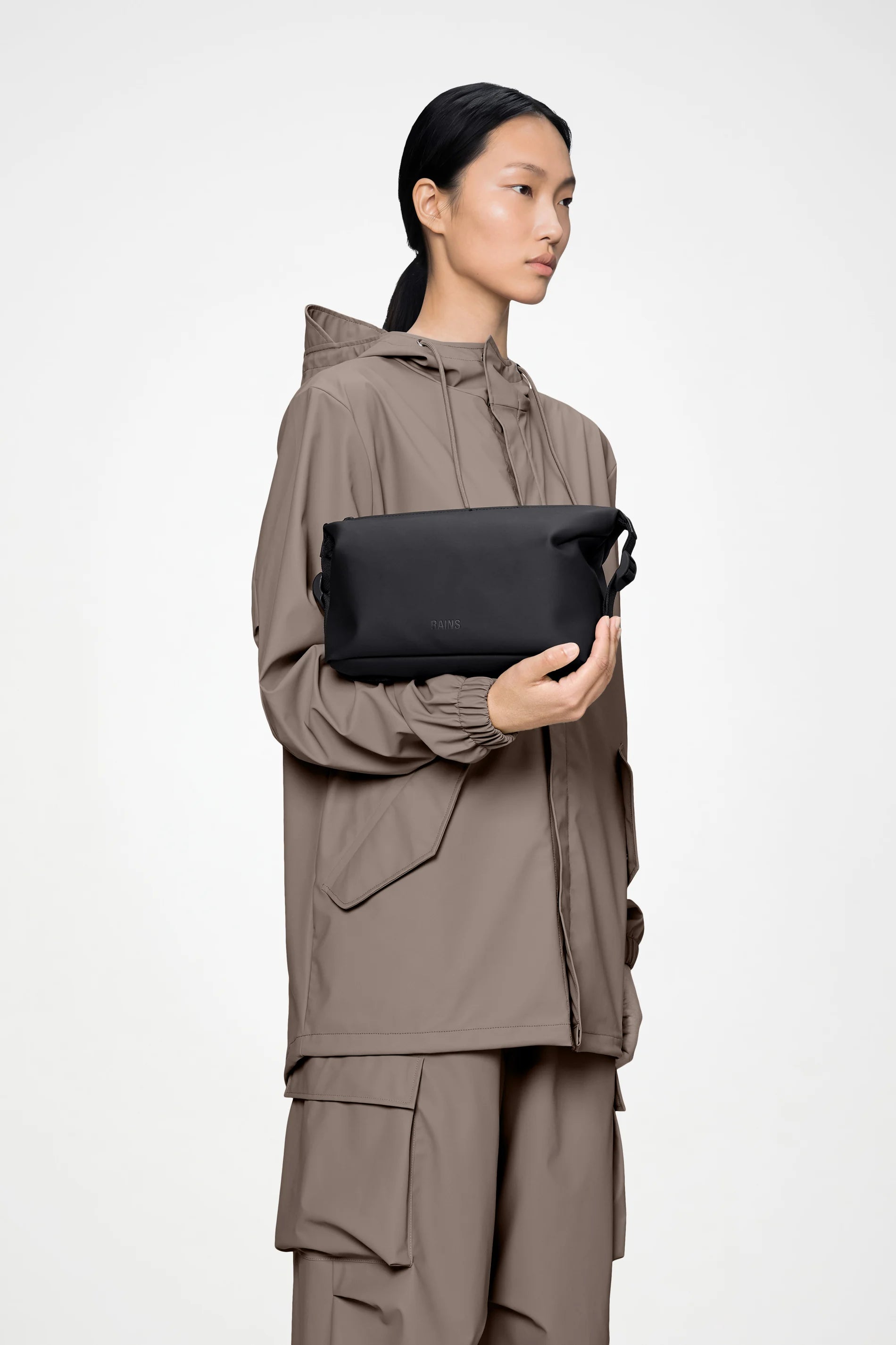 A person wearing a taupe hooded jacket and matching cargo pants holds the Rains Hilo Wash Bag W3 in Black by Rains in front of a plain light background.
