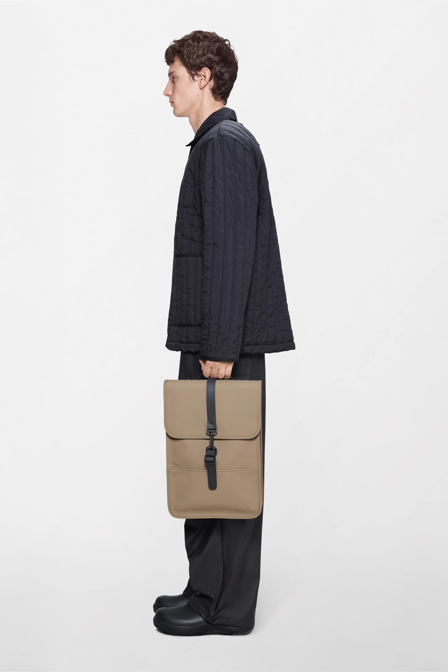 A person dressed in a black quilted jacket, black pants, and black shoes stands in profile against a plain light background, holding the Rains Backpack Mini Bag in Beige by Rains.